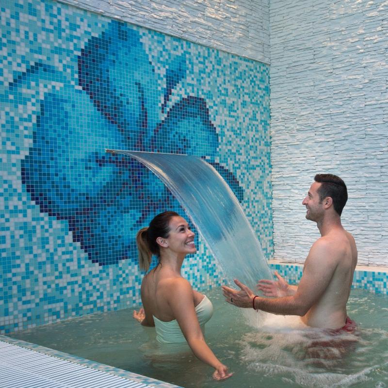 Coppia felice sotto cascata in piscina con mosaico floreale blu.